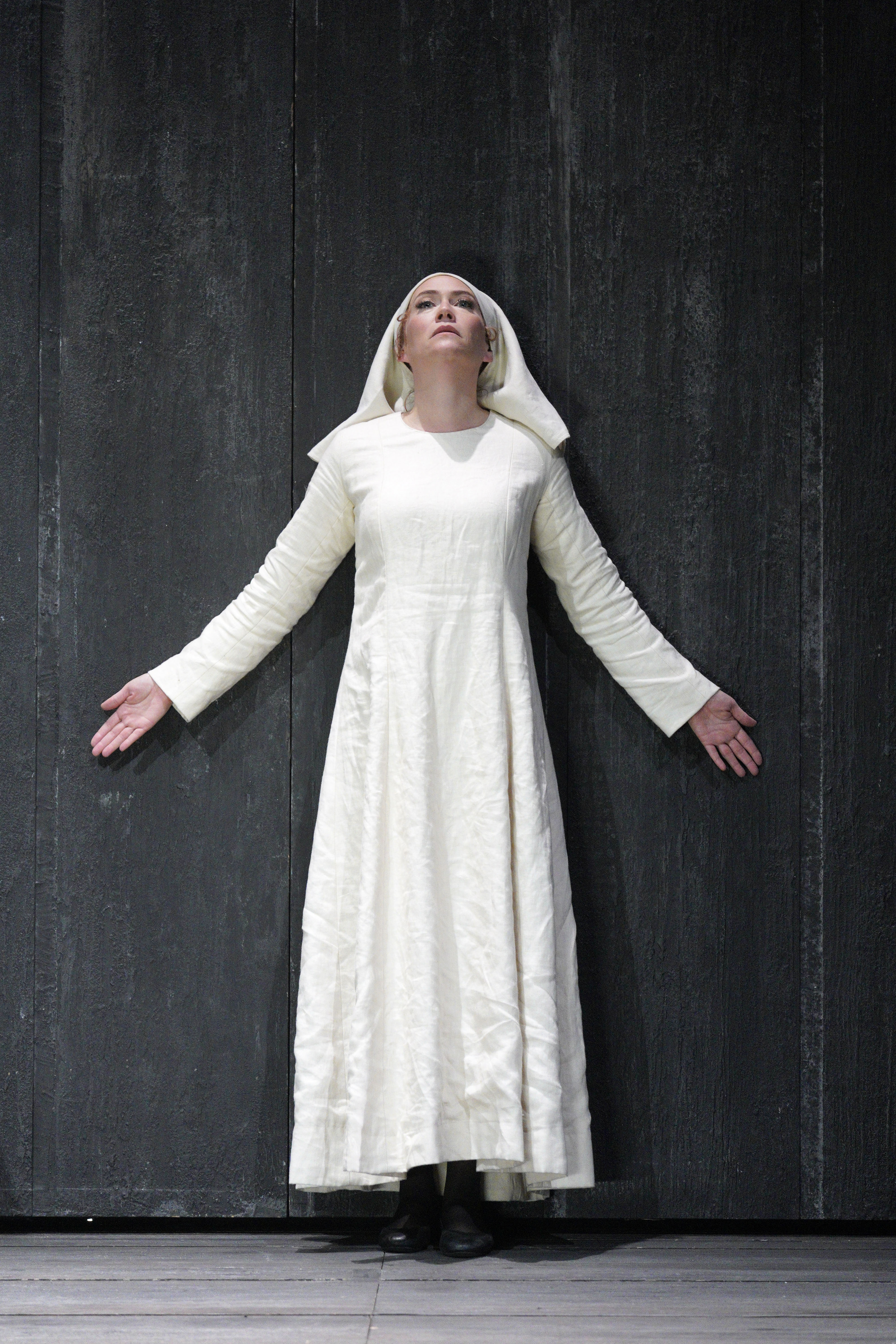 A woman in a white dress stands against a tall black wall with her arms outstretched.