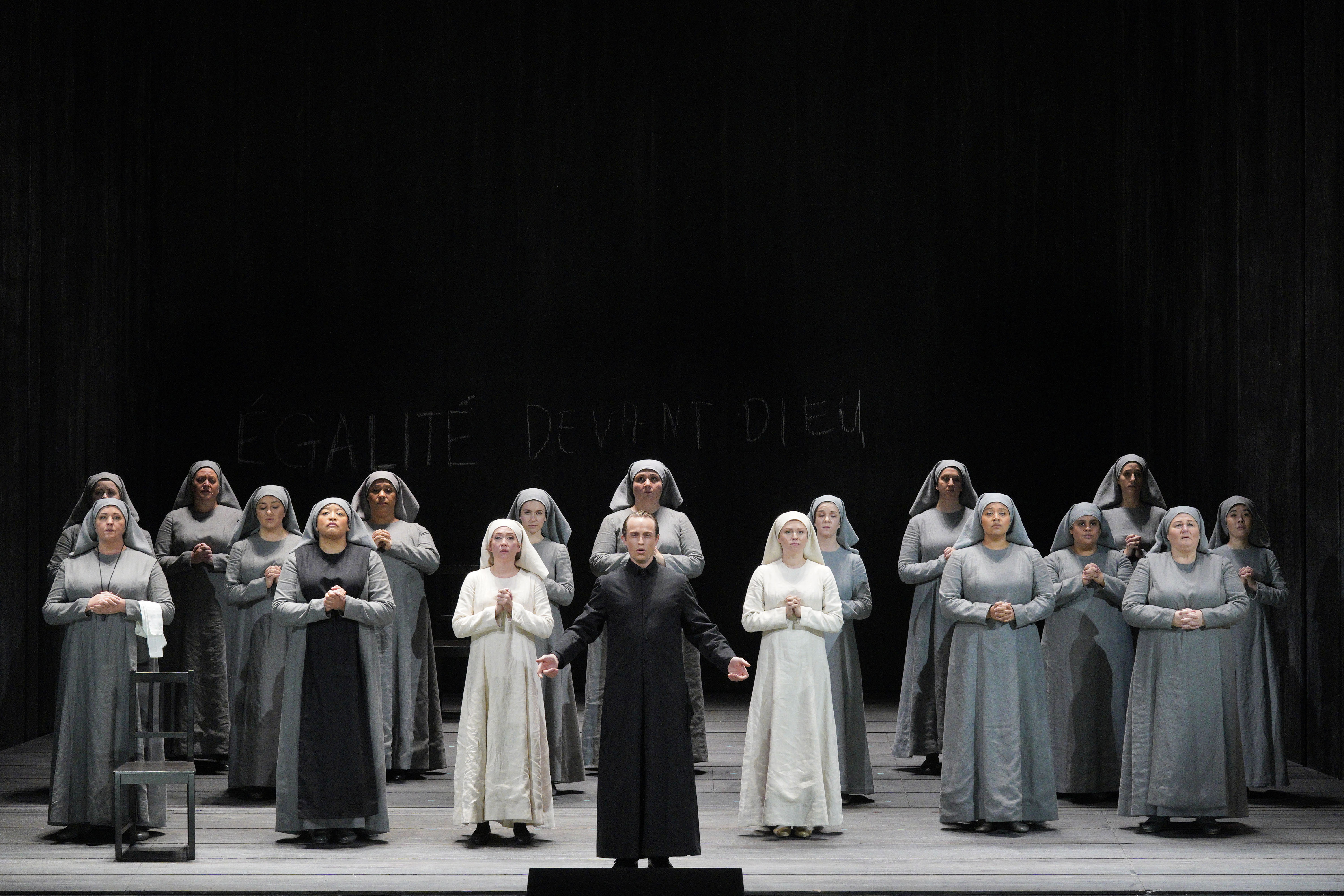 A man in black stands before rows of nuns in gray and white habits onstage.